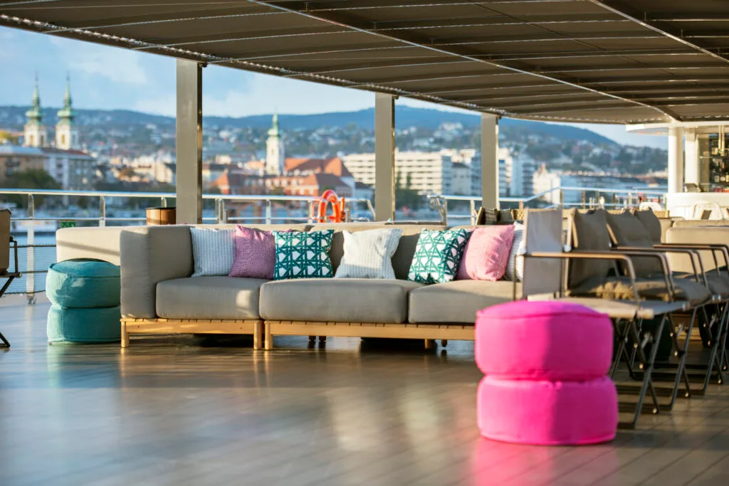 Recreational space onboard river cruise. A grey sofa with colourful cushions is next to a row of chairs.