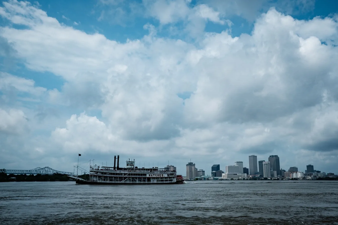 River cruise sailing alongside the horizon of a city. Tall buildings line the river. The sky is cloudy.