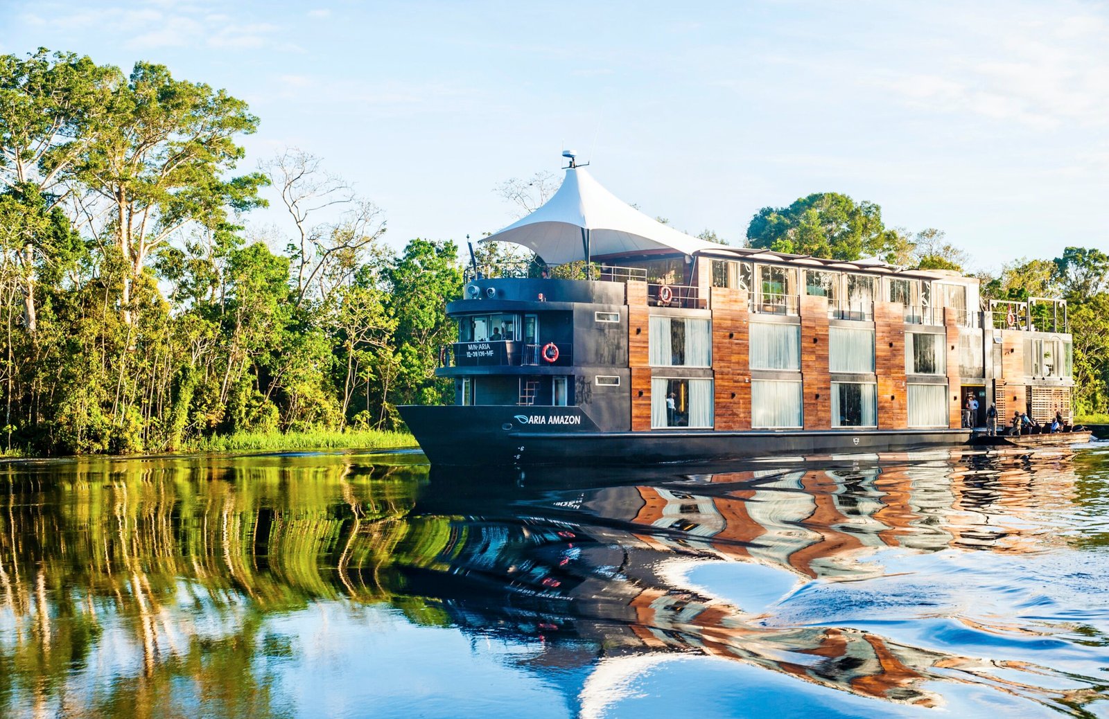 Black and wooden Aria Amazon ship sailing through glossy dark water lined with green trees.