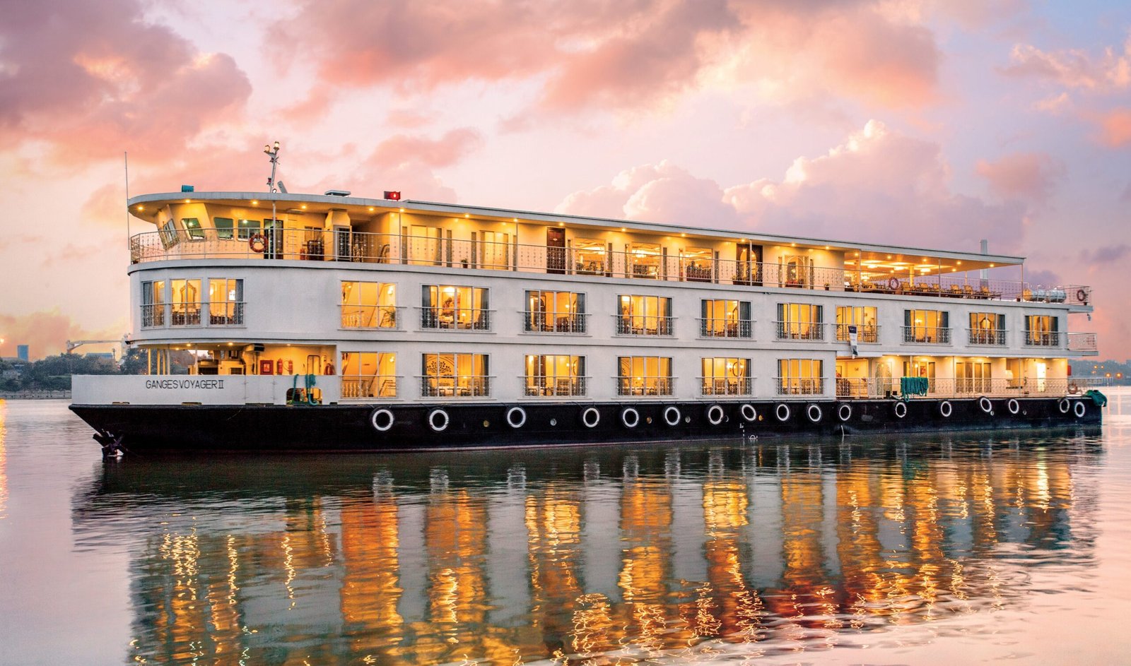 Black and white river cruise illuminated by yellow lights onboard. The sky is pink and cloudy.