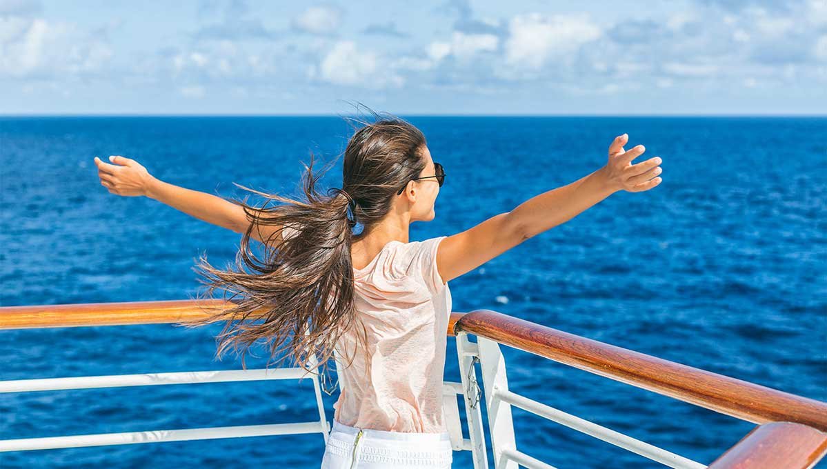 Woman with long brown hair raising her arms & smiling whilst looking out to sea from her cruise.
