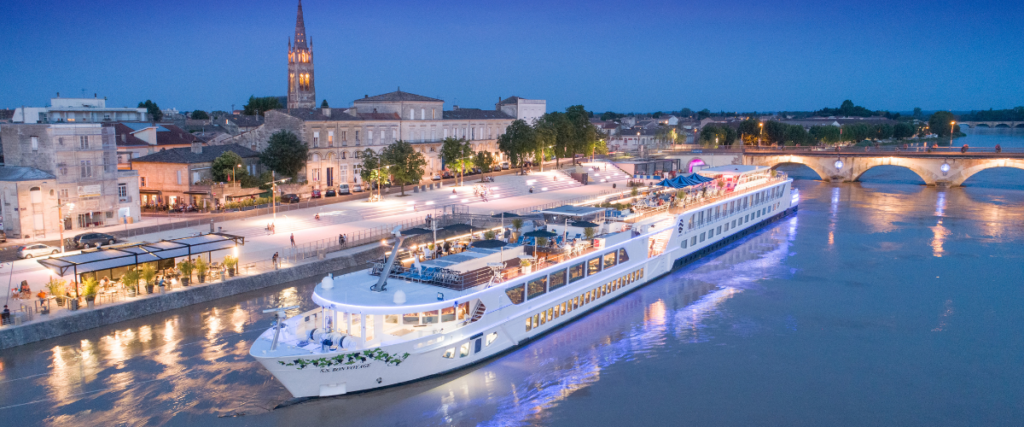 Large white river cruise sailing along side an old pretty town at night.